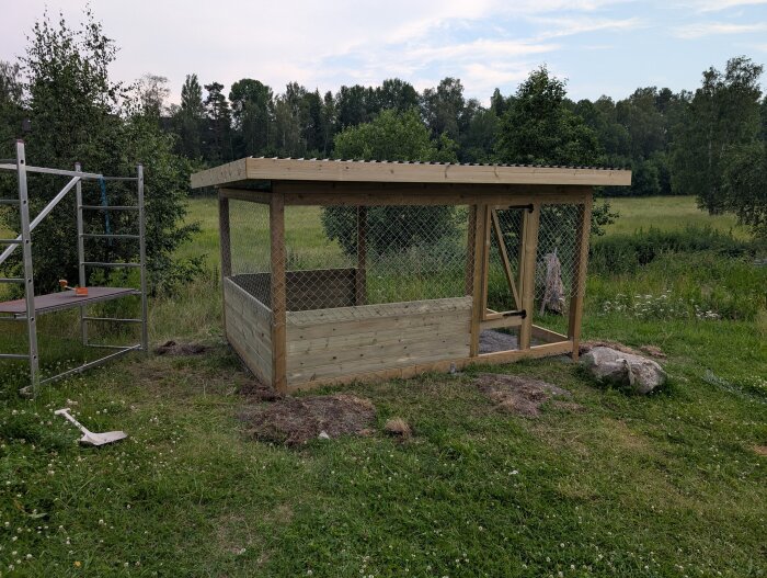 Halvfärdig kaninbur av trä och nät i trädgård med ställning bredvid, grönt gräs och skog i bakgrunden.