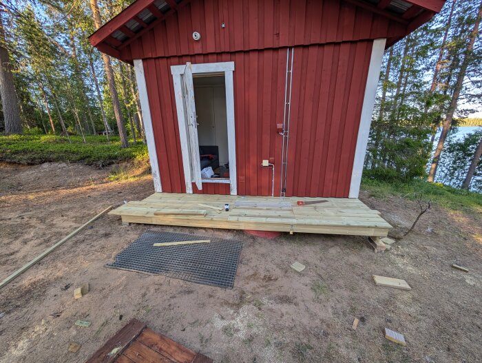 Trallprojekt framför rött hus med vit dörr och kringliggande skog. Ny trätrall installerad, omgiven av verktyg och material.