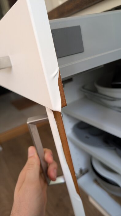 En person håller i ett kökshandtag på en vit kökslucka som delvis har lös foliering, med insida av en låda synlig i bakgrunden.