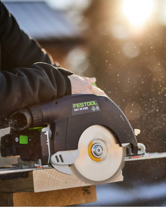 Elektrisk kapsåg Festool HKC 55 KEB sågar genom en träbit i solnedgång.