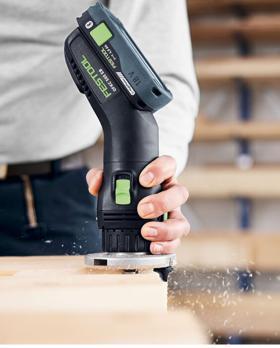 En person använder en Festool trådlös handöverfräs för att bearbeta trä, träspån flyger omkring.