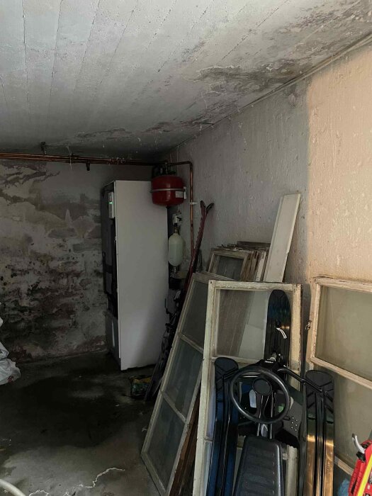 Fuktig källare med synliga vattenfläckar på väggar och golv, diverse föremål inkl. fönsterbågar och en pulka, problem med läckage från balkong.