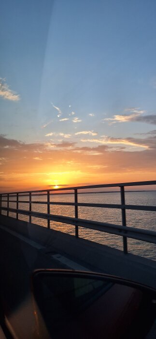 Solnedgång över havet, sedd från en bil på en bro, med orange och blå himmel och reflektioner i vattnet.