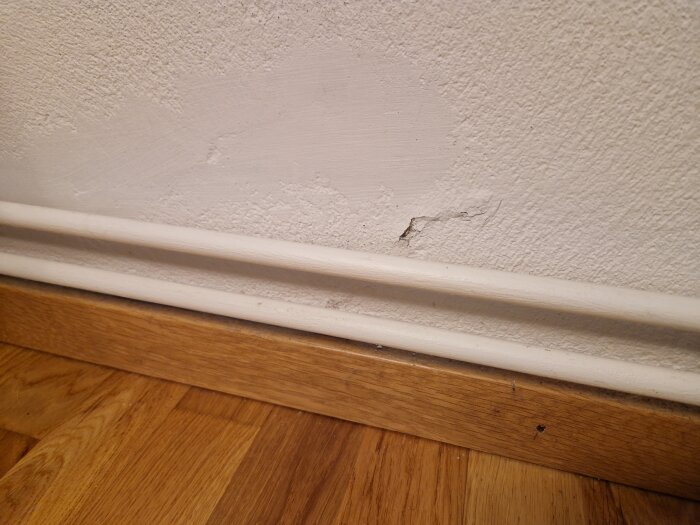 Blåsig innervägg med flagnande puts och sprickor ovanför golvlist i hus från 70-talet.