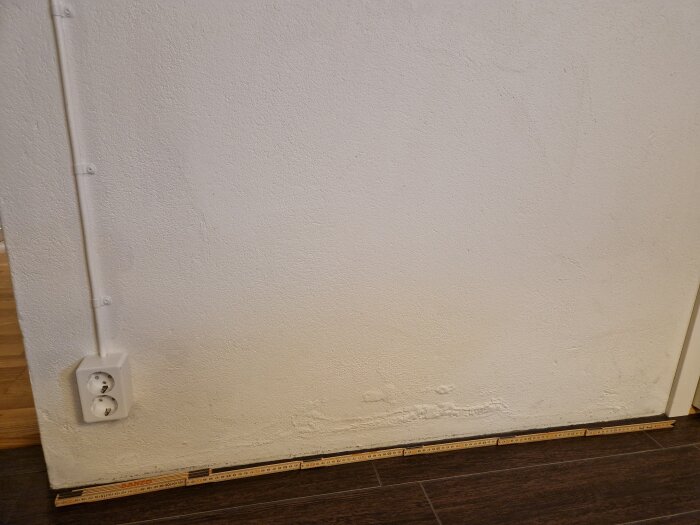 Innervägg med gulaktiga bubblor längs golvet vid en strömbrytare, bubblor verkar orsakas av fukt; golvlist och måttstock synliga.