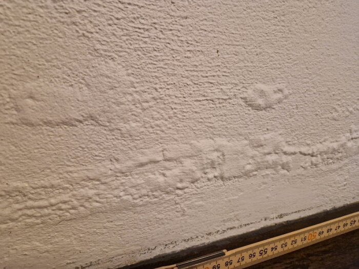Bilden visar en innervägg med bubblor och ojämnheter i putsen, samt en måttstock längs golvet för skala.