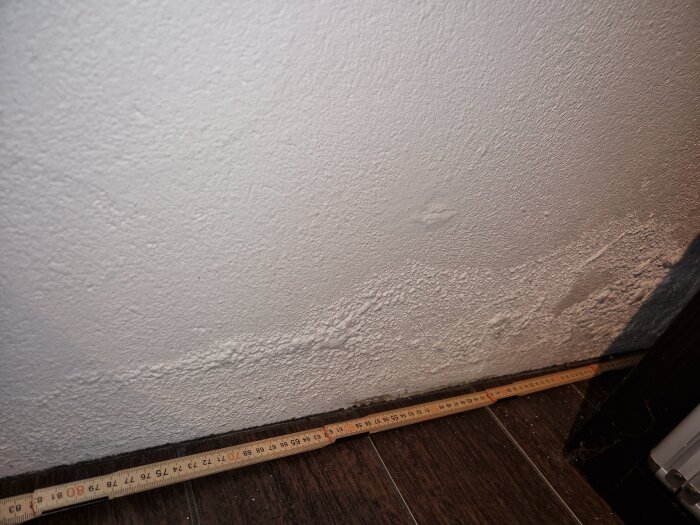 Bubbelformationer på en vit innervägg nära golvet med mätsticka nedtill, tecken på potentiella fuktproblem.