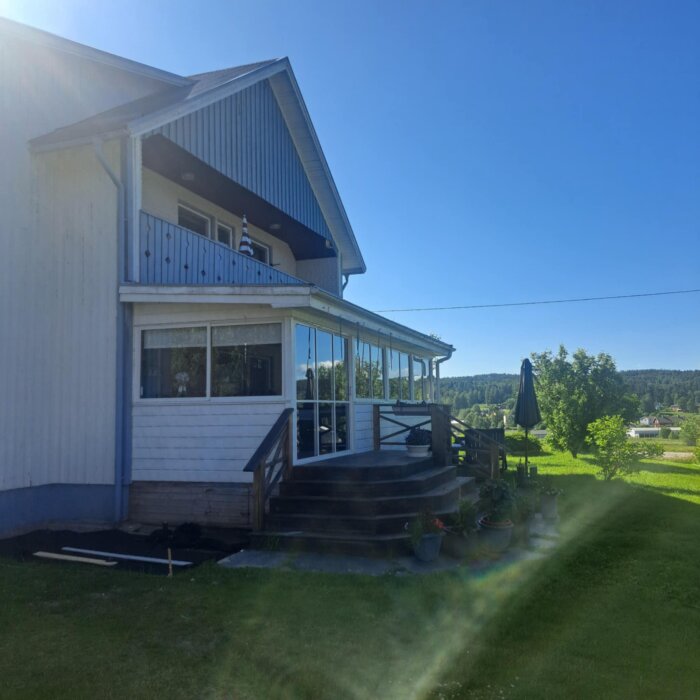 Inglasad altan på ett hus med träfasad, med trätrappa och omgivande grönområde under en klarblå himmel.