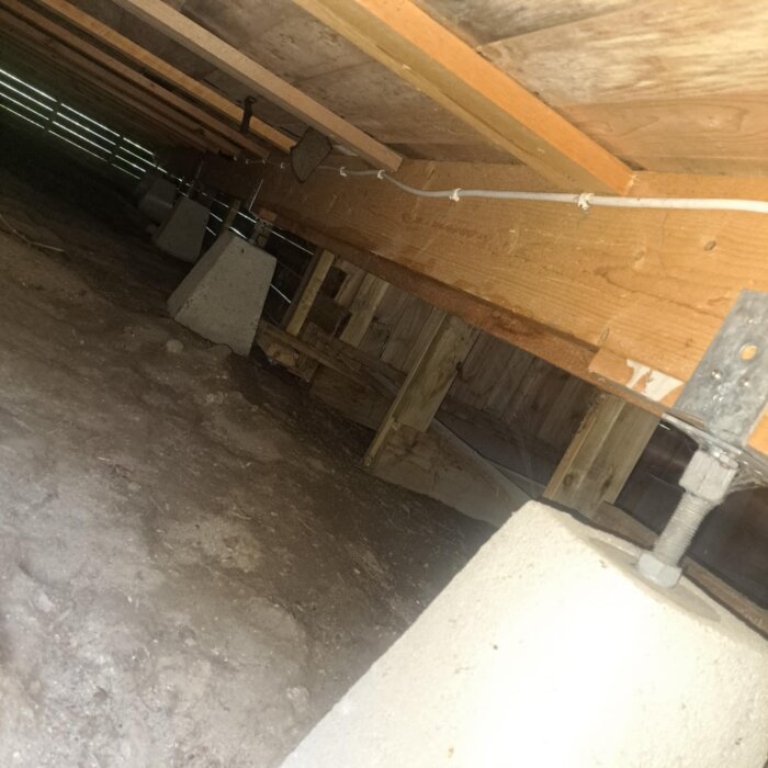 Under en altan syns träbjälkar som vilar på betongplintar över en jordyta. Ljuset strömmar in från sidan, konstruktion i trä och metall.