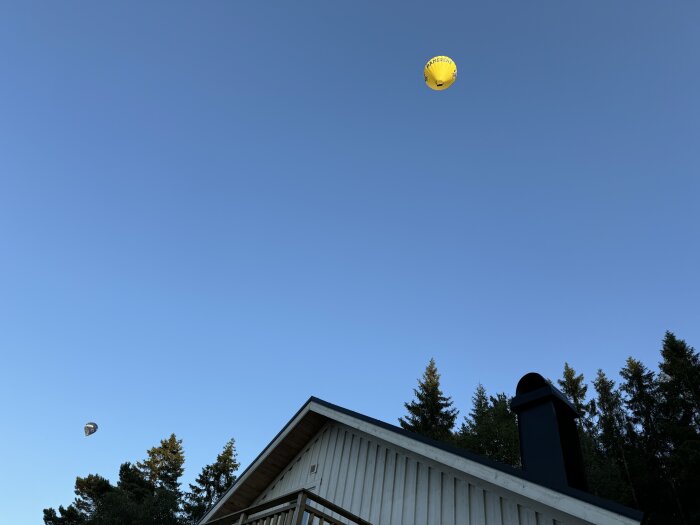 En gul och en mörk luftballong i skyn ovanför ett hus och tallar mot en klarblå himmel.