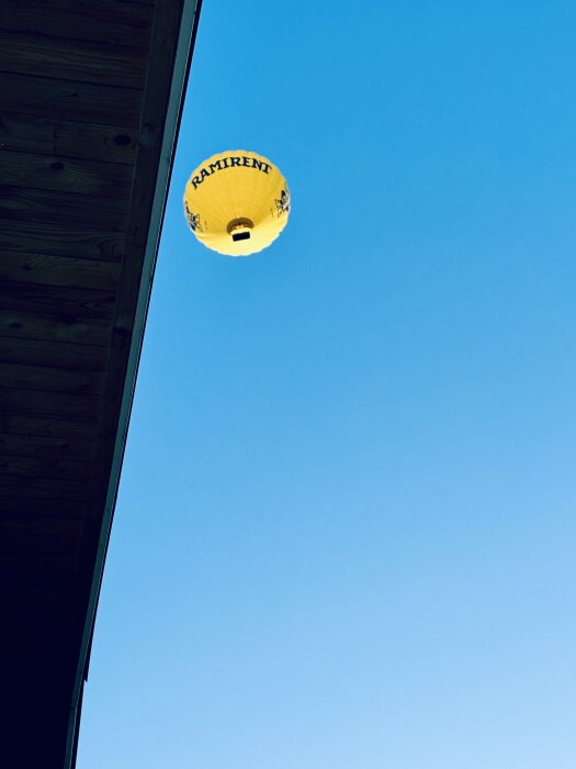 Gul luftballong med "Ramirent" svävar på en blå himmel, delvis täckt av en byggnad.