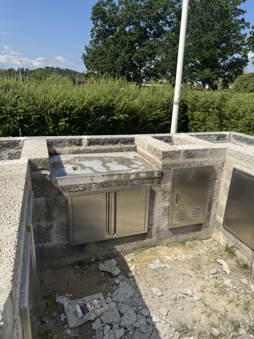 Utomhusgrill under konstruktion med nylagd skiva och plats för gjuten bänkskiva, omgiven av gröna buskar och träd.