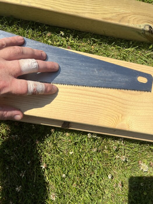 En hand med tejpade fingrar håller en fogsvans mot en träplanka på gräs.