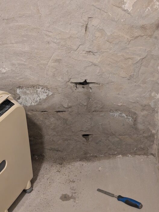 Vägg med små hål mellan blocken, bredvid syns ett beige värmeelement och ett verktyg på golvet.