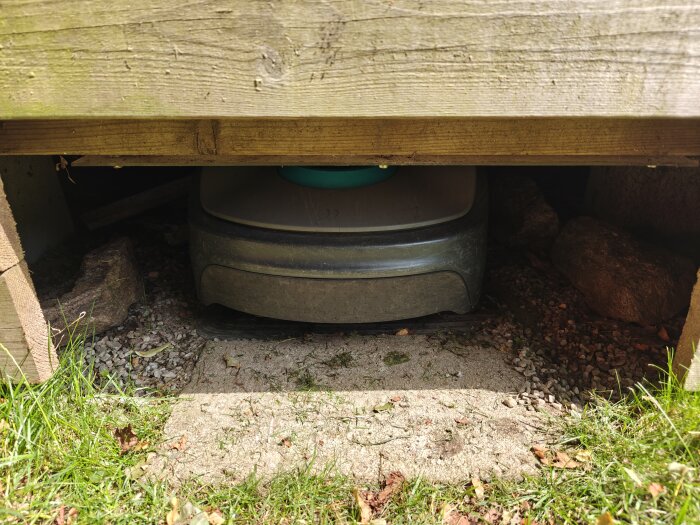 Robotgräsklippare under en träkonstruktion, dockad något snett på en stenig yta, omgiven av grönt gräs.