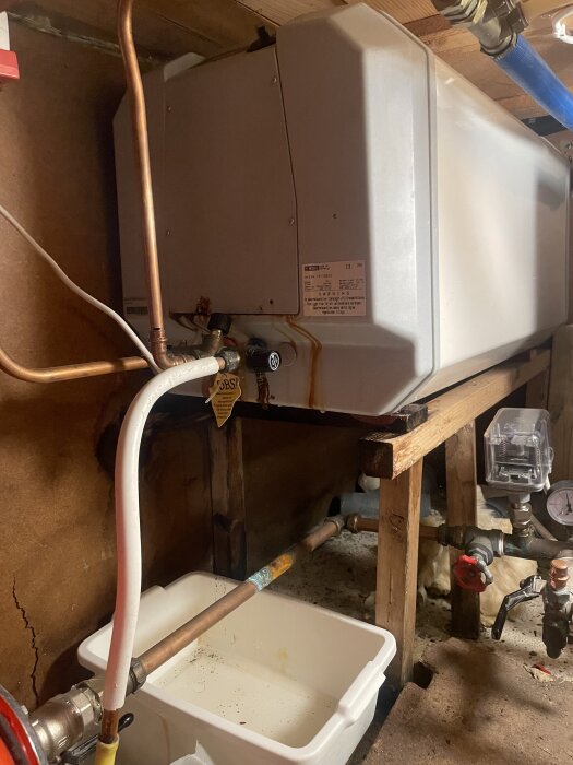 Gammal varmvattenberedare som läcker, med en vit behållare placerad under för att samla upp vatten. Synliga kopparrör och trästomme.