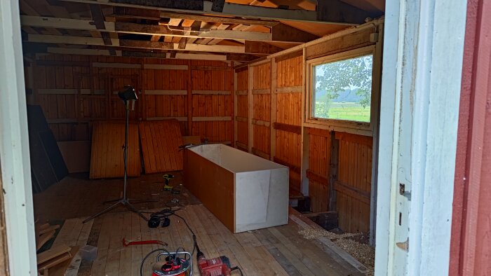 Tomt förråd med rivet trägolv, verktyg och material synliga. Ett stort fönster visar utsikt över grönt landskap.