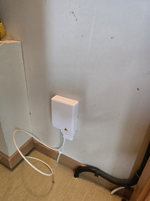 Vit väggbox monterad nära golvet med anslutna vita och svarta kablar, samt en lila fiberkabel i processen att installeras.