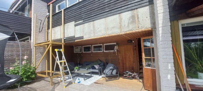 Trähusfasad under renovering med synlig betong bakom panelen, stege och verktyg i trädgården, samt en inglasad dörr.