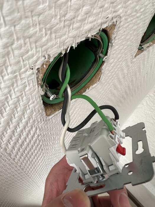 Eluttag och tre kablar, svart, vit och grön, i en grön infälld dosa i väggen, hålls av en hand för installation.