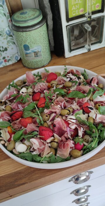 Sallad med skinka, jordgubbar, oliver och gröna blad i en vit skål på en träbänk, omgiven av färgglad burk och teförpackningar.