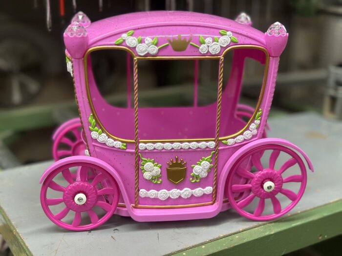 En rosa leksaksvagn formad som en prinsessvagn med dekorationer av vita blommor och kronor, står på ett bord i en verkstad.