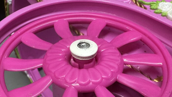 Ett rosa plasthjul med en vit mutter i centrum, en del av en barnleksak som behöver repareras.