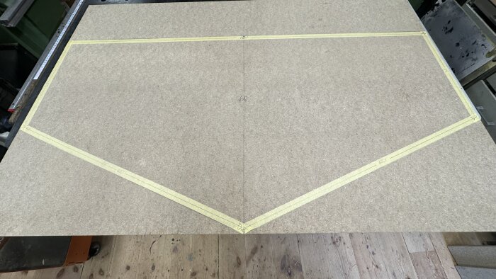 Spånskiva med inritad form för spelplan, markerad med måttband och tejpade linjer, ligger på arbetsbänk i träverkstad.