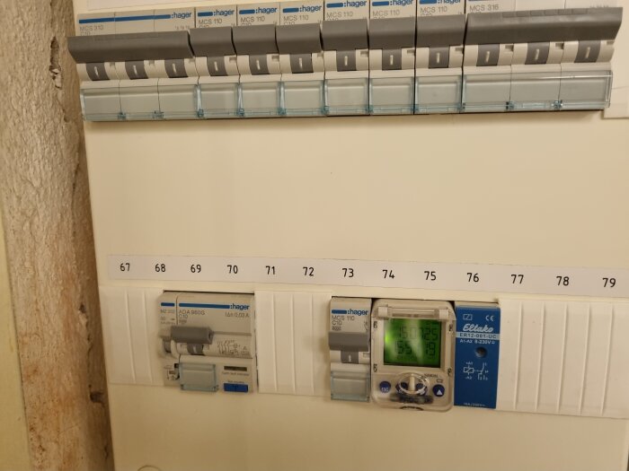 Elcentral med automatsäkringar och en digital display, numrerade från 67 till 79, på en vägg med ojämn yta.