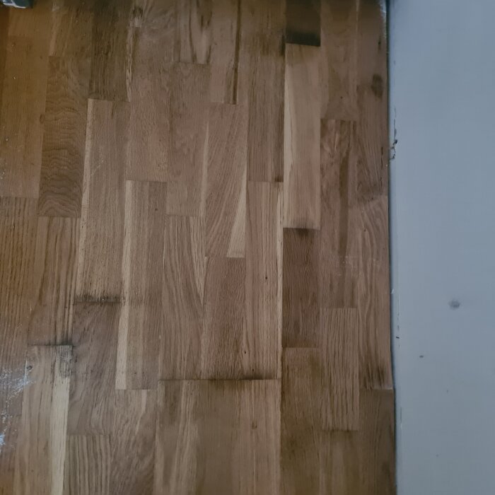 Trägolv med mörka fläckar och eventuell vattenskada nära en vägg, orsakat av vattenläckage från ett kylskåp.