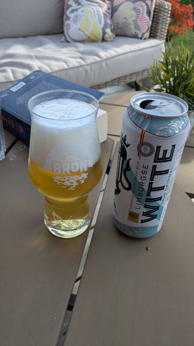 Ett glas öl med skum och en burk öl märkt "Limburgse Witte" står på ett bord utomhus, med en bok och en soffa i bakgrunden.