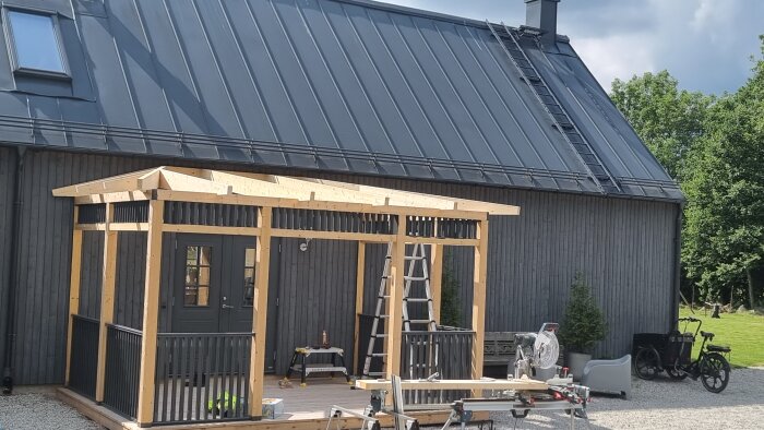 Träpergola under konstruktion framför ett gråmålat hus med sadeltak. Stege och verktyg syns i förgrunden, grönska i bakgrunden.