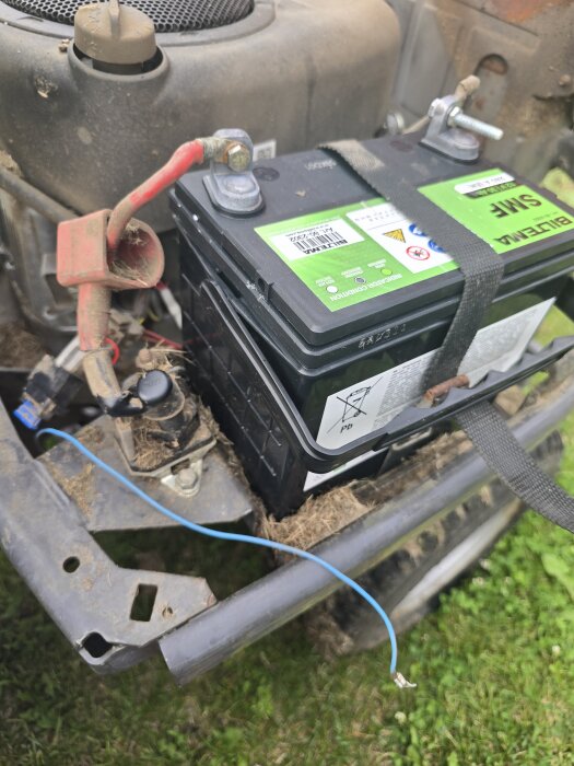 En Husqvarna R216 gräsklippares batteri med en lös blå sladd synlig framför maskinen.