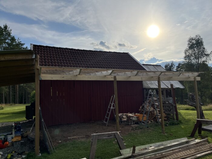 Träpergola under uppförande bredvid röd ladugård, med stege och ved i bakgrunden, soligt väder syns på himlen.