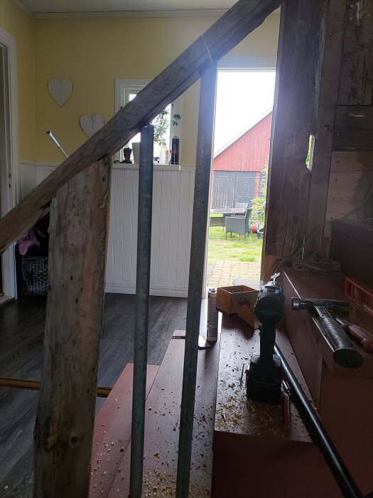 Trappa under renovering med verktyg och träspån. Utanför syns en röd lada och trädgårdsmöbler.