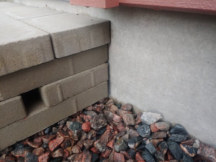 Hörn av entrétrappa med mur- och markstenar nära husgrund, omgiven av småsten.