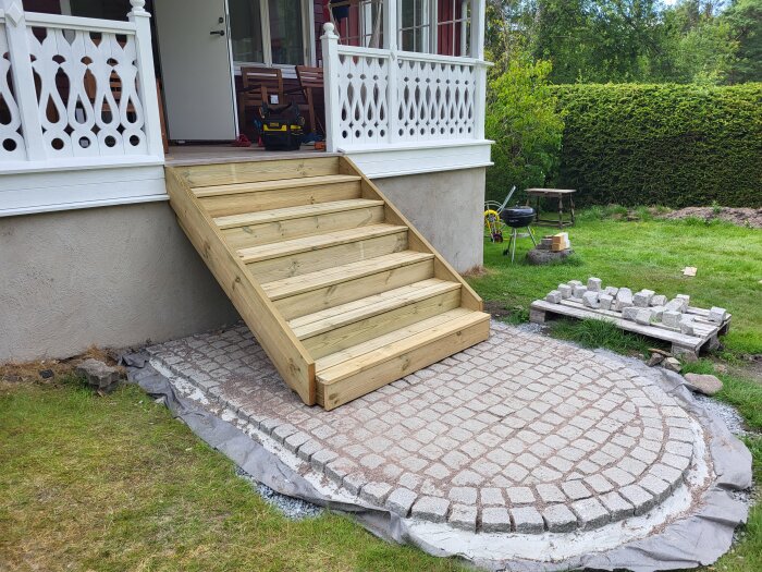 Nybyggd trätrappa leder från en veranda ner till en stenlagd yta i en trädgård.