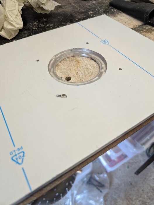 Fräst aluminiumplatta med cirkulärt urtag, omgiven av arbetsyta med handskar och verktyg.