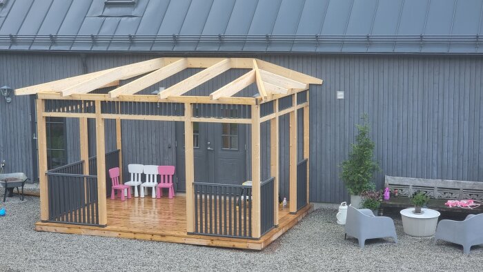 Trästomme för pergola under byggnation med trädgårdsmöbler och barnstolar framöver ett grått hus. Ljust trä och svart detaljer.