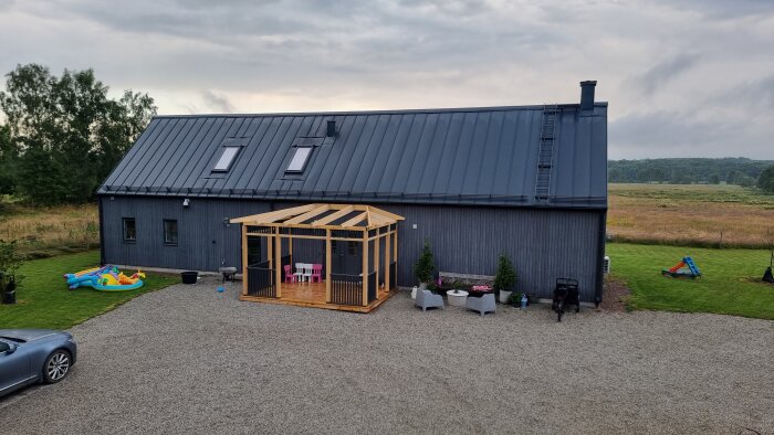 Ett grått hus med svart tak, en träpergola och en uppblåsbar barnpool i trädgården, omgiven av gröna fält och molnig himmel.