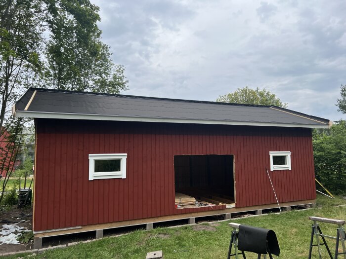 Rött träskjul under konstruktion med tak täckt av filt, två fönster och en stor rektangulär öppning, står på plintar i en grön trädgård.