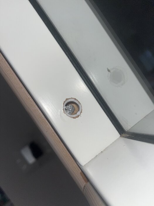 Närbild på en vit fönsterkarm med en borrad skruv i träet, omgiven av spår av färg och trä.