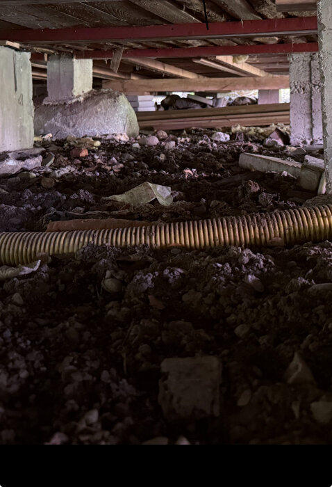 Öppen plintgrund under ett fritidshus med synlig jord och en dräneringsslang, samt träreglar ovanför.