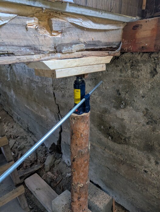 Domkraft lyfter hus med 45x220 reglar som stöd. Ett trästycke och järnrör används för att sprida vikt. Markens stabilitet visas vid en sprucken grundmur.