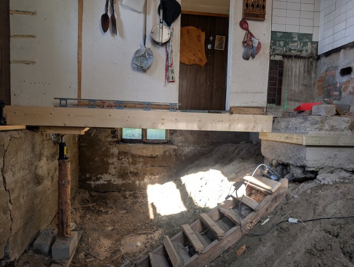 Byggprojekt där en husgrund har lyfts med träbjälkar, underlag av trä, betongblock och en domkraft synlig under huset.