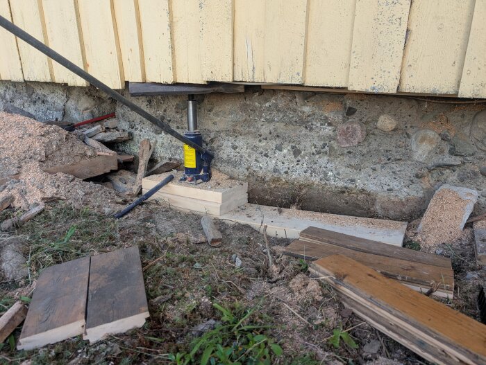 Husets grund renoveras med hjälp av en domkraft. Träplank och sågspån syns runt omkring.