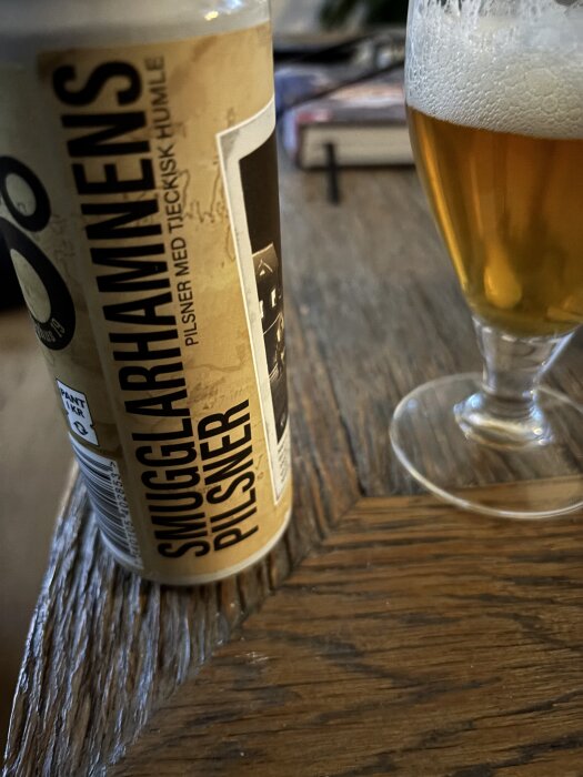 En burk Smugglarhamnens pilsner med en fylld ölglas på träbord.