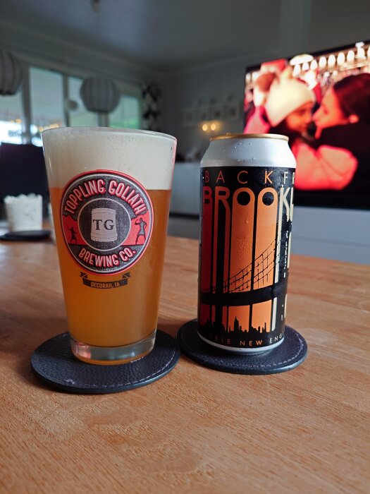 En öl från Toppling Goliath i ett glas bredvid en burk öl på ett bord framför en tv.