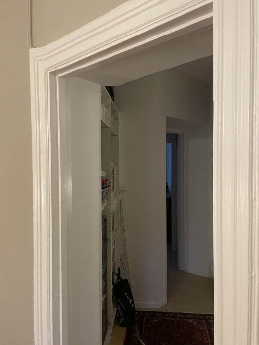Öppning utan dörr in i ett rum, visar dörrkarm utan gångjärn. I bakgrunden syns hyllor och en mörkare dörröppning. Målade ytor och ljusa väggar.