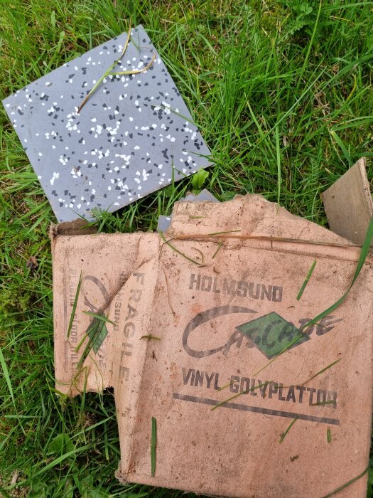 Förpackning av vinylgolvplattor med text "Cascade" och en grå golvplatta med vitt och svart mönster ligger på gräs.